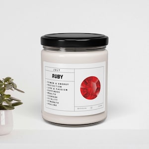 May include: A white candle jar with a black lid and a red gemstone image. The label reads "JULY RUBY" and lists the gemstone's properties, including "power & energy", "protection", "love & passion", "happiness", "wealth", "courage", "vitality", "strength", and "healing".