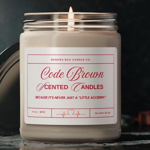 May include: A brown glass jar candle with a white label that reads "Code Brown Scented Candles" and "Because it's never just a "little accident."". The label also includes the company name "Banana Bag Candle Co.", a heart rate graphic, and the text "3 oz. NPO" and "Do Not Drink".