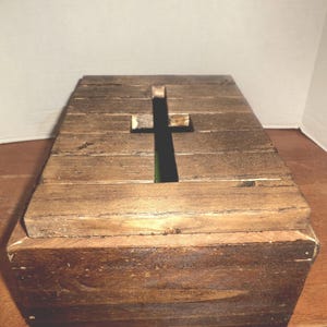 May include: A wooden box with a cross-shaped opening on the top. The box is constructed from planks of wood, with a natural, aged appearance. The cross cutout reveals a dark interior.