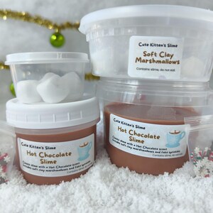 May include: A stack of clear plastic containers filled with slime and soft clay marshmallows. The containers have white lids and labels that read "Cute Kitten's Slime" and "Hot Chocolate Slime." Small bags of decorative sprinkles are also included.