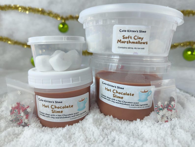 May include: A stack of clear plastic containers filled with slime and soft clay marshmallows. The containers have white lids and labels that read "Cute Kitten's Slime" and "Hot Chocolate Slime." Small bags of decorative sprinkles are also included.