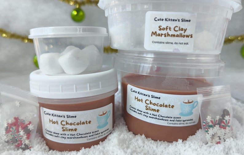 May include: Several containers of "Cute Kitten's Slime" are displayed, including a jar of brown "Hot Chocolate Slime" and a container of white "Soft Clay Marshmallows." The labels include a hot chocolate cup graphic and the text "Contains slime, do not eat."