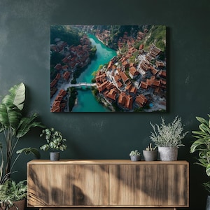 Puede incluir: Vista aérea de un pueblo con tejados de tejas rojas junto a un río turquesa. Un puente blanco cruza el agua, conectando el pueblo. El paisaje circundante presenta verdes colinas, creando una escena pintoresca.