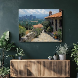 May include: A framed canvas print depicting a scenic landscape. A stone path leads to a building with a red tile roof. The background features mountains and a blue sky with clouds. Green foliage and flowers border the path and building.