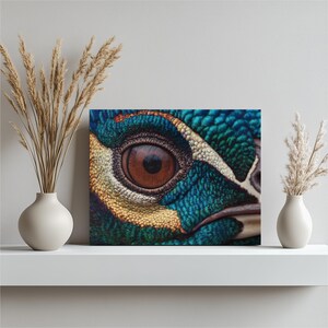 Puede incluir: Primer plano de una impresión en lienzo que presenta el ojo detallado de un ave. El ojo es de un marrón intenso, rodeado de vibrantes plumas azules, verdes, amarillas y blancas. La obra de arte se exhibe en una estantería blanca.