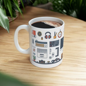 May include: White ceramic coffee mug filled with dark coffee, featuring a repeating pattern of tech-related icons in various colors. The icons include headphones, keyboards, and computer monitors. The mug sits on a light wood surface.