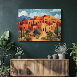 May include: A watercolor painting depicts a desert village with orange and yellow buildings under a blue sky with clouds. Green trees and cacti dot the landscape, with a mountain in the background. The artwork is displayed on a wall.