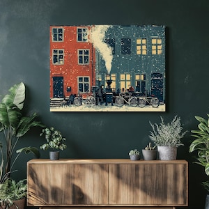 May include: A canvas print depicts a snowy street scene with red and blue buildings. People and bicycles are visible in the snow. A plume of smoke rises from a chimney, and the windows glow with warm light.