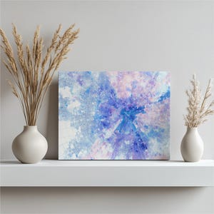 May include: An abstract watercolour canvas print in shades of blue and purple. The artwork is displayed on a white shelf, with two white vases holding dried ornamental grass on either side.