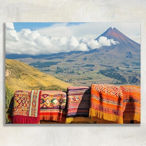 May include: A scenic landscape featuring a mountain with a red peak under a cloudy sky. In the foreground, colorful woven textiles with fringe hang, showcasing intricate patterns in red, orange, and purple.