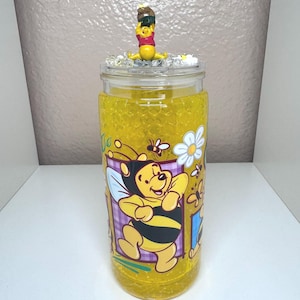 May include: A glass jar filled with yellow liquid and decorated with a Winnie the Pooh theme. The jar features a cartoon image of Winnie the Pooh and bees. The lid is adorned with a Winnie the Pooh figurine. The jar is approximately 6 inches tall.