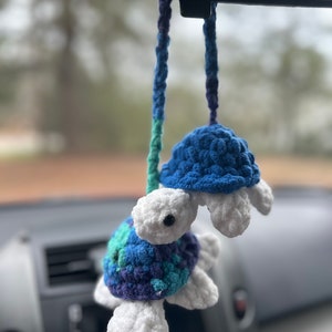 May include: Two crocheted turtle car air fresheners hanging from the rear view mirror of a car. The top turtle is blue and the bottom turtle is purple, blue, and green.