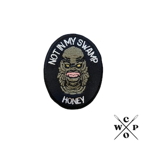 May include: Black and white embroidered patch featuring a green creature with yellow eyes and a pink mouth. The text on the patch reads "Not in my swamp Honey".