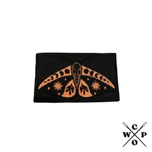 May include: A black embroidered patch featuring a moth with orange wings and black details. The moth has a stylized design with crescent moons and stars on its wings.
