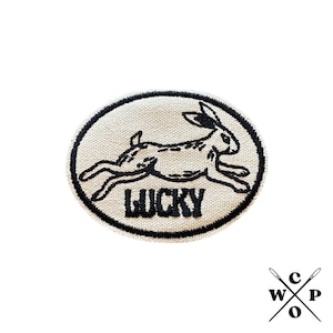 Op de afbeelding: Een witte ronde patch met een zwarte rand. De patch heeft een zwart geborduurd beeld van een lopend konijn met het woord "LUCKY" eronder.