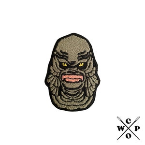 May include: Embroidered patch featuring the Creature from the Black Lagoon. The patch is green and black with yellow eyes and a pink mouth.