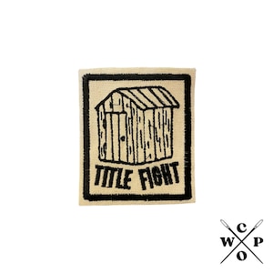 以下が含まれることがあります： 小さな家と「TITLE FIGHT」という文字が刺繍された黒と白のワッペンです。