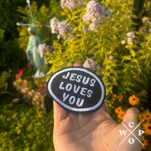 May include: A black and white embroidered patch with the text "JESUS LOVES YOU" in a white oval on a black background.