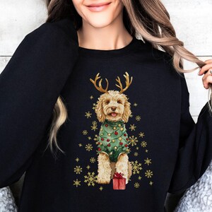 May include: Black sweatshirt with a watercolour illustration of a dog dressed as a Christmas tree, complete with antlers and a red gift. Gold snowflakes surround the dog. A festive holiday design.