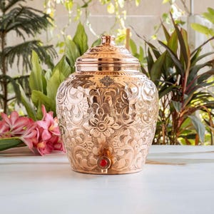 May include: A polished copper water dispenser featuring an intricate embossed floral and scroll pattern across its rounded body. The vessel includes a matching lid and a small, circular red-accented spigot at the base for dispensing liquid.