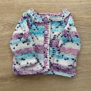 Peut inclure: Un cardigan pour bébé tricoté à la main, aux couleurs blanc, violet et bleu. Le pull présente un col festonné, une fermeture à boutons et un motif rayé. Le vêtement est en fil doux et est conçu pour un bébé.