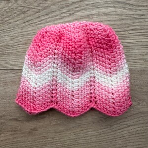 May include: A hand-knitted baby hat with a scalloped edge. The hat has a chevron pattern in shades of pink and white. The hat is made from soft yarn and is designed for a baby.