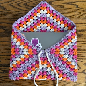 Puede incluir: Una funda para portátil de ganchillo hecha a mano y colorida. La funda presenta un diseño de cuadrados de la abuela en tonos morado, rosa, naranja y blanco. Tiene un cierre de cordón y está abierta, revelando un portátil con una pegatina.