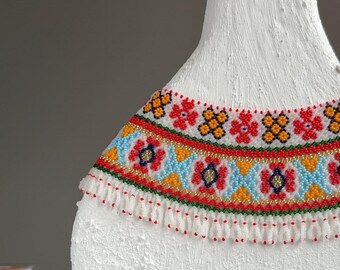 Może przedstawiać: Biały, teksturowany obiekt z kolorowym kołnierzem z koralików. Kołnierz ma powtarzający się wzór czerwonych, żółtych, niebieskich i złotych koralików. Dolna krawędź kołnierza ma białe frędzle.