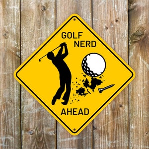Puede incluir: Señal amarilla en forma de diamante con texto negro y una silueta de un golfista. La señal dice "Golf Nerd Ahead" con una bola de golf explotando en pedazos.