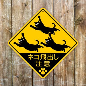 Könnte beinhalten: Gelbes, rautenförmiges Schild mit schwarzen Katzenillustrationen und japanischem Text. Das Schild zeigt drei schwarze Katzensilhouetten mit Masken, mit dem japanischen Text "Neko Tobidashi Chui" und einem Pfotenabdrucksymbol.