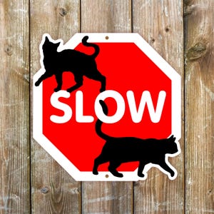 May include: An octagonal red sign with a white border and the word SLOW in large white capital letters. Two black cat silhouettes are positioned on the sign, one walking across the top and one walking across the bottom section.