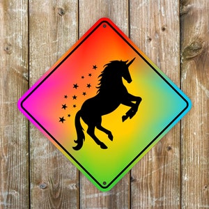 May include: A diamond-shaped sign with a rainbow gradient background featuring a black silhouette of a rearing unicorn and stars. The sign has a black border and is mounted on a wooden background.