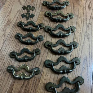 Puede incluir: Un conjunto de tiradores y bocallaves de latón antiguo dispuestos sobre una superficie de madera. Los tiradores tienen un diseño curvo con detalles decorativos. Las bocallaves son pequeñas y ornamentadas.