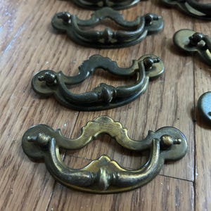 Op de afbeelding: Een verzameling antieke messing lade-grepen met een gebogen handvatontwerp. De grepen hebben een vintage esthetiek, met een aangetaste afwerking en sierlijke details. De hardware is gerangschikt op een houten oppervlak.