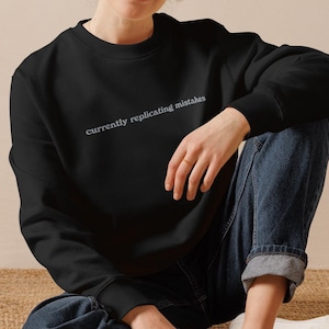 Könnte beinhalten: Schwarzes Sweatshirt mit dem Text "currently replicating mistakes" in heller Farbe. Das Sweatshirt wird mit blauen Jeans und weißen Turnschuhen kombiniert. Die Person sitzt auf einer gewebten Oberfläche.