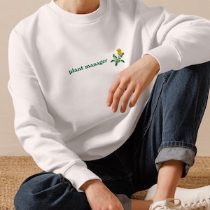 Könnte beinhalten: Ein weißes Sweatshirt mit dem grünen Schriftzug "plant manager" und einer kleinen gelben Blumen-Grafik. Das Sweatshirt wird mit blauen Jeans und weißen Turnschuhen kombiniert. Die Person sitzt auf einer gewebten Matte.