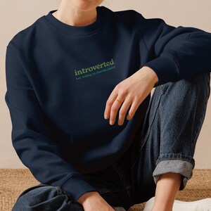 Könnte beinhalten: Marineblaues Sweatshirt mit dem Wort "introverted" in grüner Schrift. Das Sweatshirt hat einen Rundhalsausschnitt und lange Ärmel. Der Text "the voice of an introvert speaks" steht unter dem Wort "introverted".