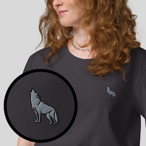 Pode incluir: T-shirt cinzento escuro com um pequeno bordado de um lobo a uivar no peito. A t-shirt é de cor lisa e tem um decote redondo clássico. Uma imagem detalhada do lobo é mostrada numa inserção circular. Várias opções de cores são exibidas.