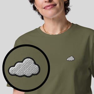 Könnte beinhalten: Olivgrünes T-Shirt mit einem kleinen, gestickten Wolkenmotiv auf der Brust. Die Wolke ist weiß mit schwarzer Umrandung. Das Bild zeigt auch eine Farbpalette der verfügbaren T-Shirt-Farben.