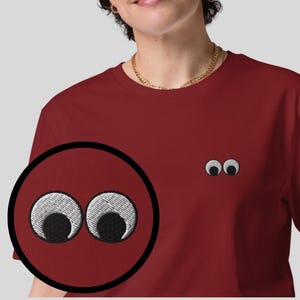 Può includere: Una maglietta color granata con un paio di occhi ricamati bianchi sul davanti. Gli occhi sono grandi e hanno pupille nere.