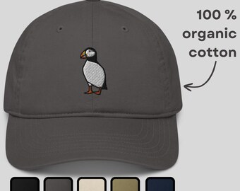 May include: A charcoal gray baseball cap embroidered with a puffin bird. The cap is made of 100% organic cotton and features a curved brim. The image also shows a selection of color options: black, charcoal, cream, olive, and navy.