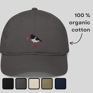 Puede incluir: Gorra de béisbol gris carbón con un diseño de pájaro bordado. El pájaro es negro, blanco, rojo y amarillo. El texto "100% organic cotton" está impreso en el lado derecho de la gorra. La gorra tiene una visera curva y un botón en la parte superior.