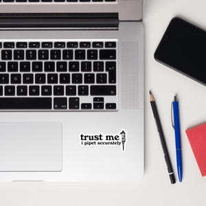 May include: A white sticker with the text "trust me i pipet accurately" in black, along with a pipette illustration. The sticker is on a laptop, with a black pencil, blue pen, and a red notepad nearby.