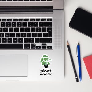 Può includere: Un adesivo con una pianta verde in un vaso nero e la scritta "plant manager" in nero. L'adesivo è su una superficie bianca, accanto a un laptop, una matita nera, una penna blu e un blocco note rosso.