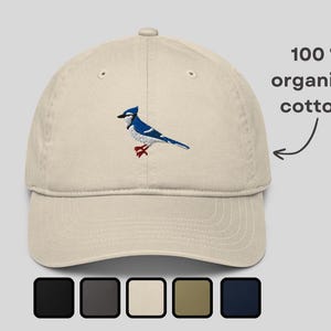 Könnte beinhalten: Beige Baseballkappe mit gesticktem Blauhäher-Design. Der Vogel ist blau und weiß mit roten Füßen. Der Text "100% organic cotton" ist abgebildet. Das Bild zeigt auch Farboptionen für die Kappe.