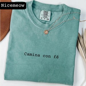 May include: A seafoam green Comfort Colors t-shirt with the text "Camina con fé" in black. A silver necklace with a small pendant rests on the shirt. The shirt has a crew neck and short sleeves.