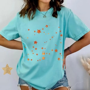 Puede incluir: Una camiseta azul claro con una constelación de estrellas de purpurina naranja. Las estrellas varían en tamaño y están dispuestas en un patrón fluido. La camiseta tiene mangas cortas y un corte holgado.