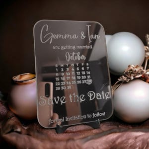 Op de afbeelding: Een heldere acryl Save the Date kaart met de namen "Gemma & Ian" en de maand "Oktober" gegraveerd. De kaart bevat een kalender en de tekst "Save the Date" en "formal Invitation to follow".