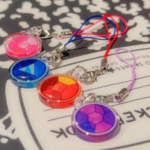 May include: Four circular acrylic charms featuring geometric gemstone patterns in pink, blue, red, and purple. Each charm includes a silver-toned metal lobster clasp and a thin nylon cord in a matching color, designed for attachment to accessories.