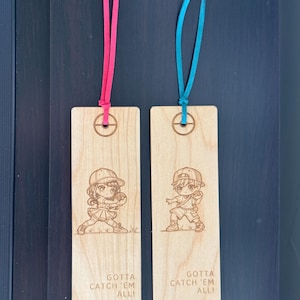 May include: Two wooden bookmarks with a cartoon illustration of a boy and girl holding a Poké Ball. The bookmarks are engraved with the text "Gotta Catch 'Em All!" and have a red and blue ribbon attached.